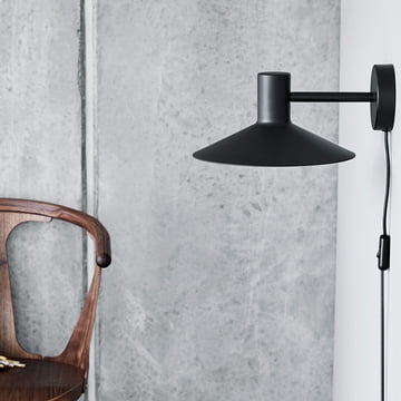 De Minneapolis wandlamp van Frandsen in het zwart op een grijze muur naast een donkere houten stoel.