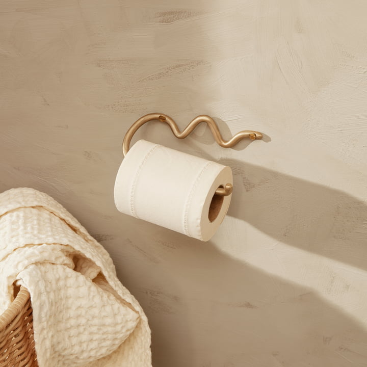 De Curvature toiletpapierhouder van ferm Living op een lichte stenen muur naast een rotan mand