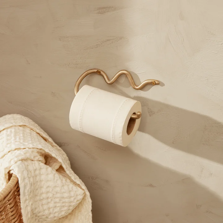 De Curvature toiletpapierhouder van Ferm Living op een lichte stenen muur naast een rotan mand