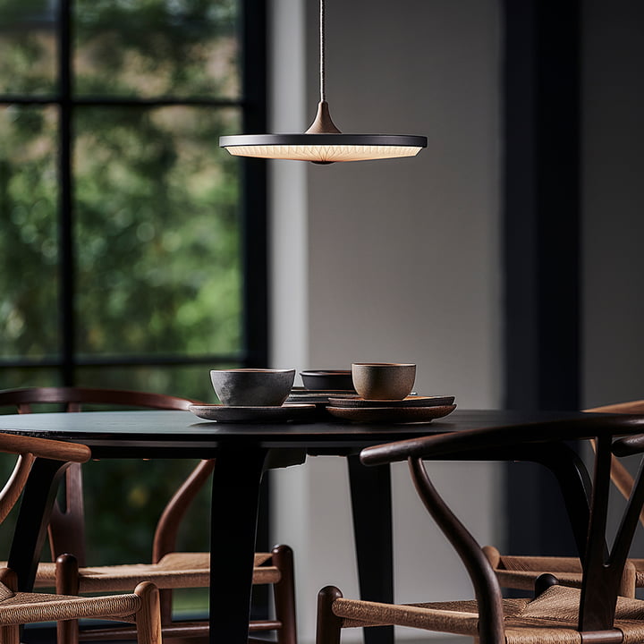 De Soleil LED-hanglamp van Le Klint boven een donkere, ronde eettafel met houten stoelen.